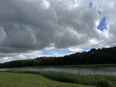 Dramatic storm clouds over river shoreline with dense trees, calm water, reed Stock Photos