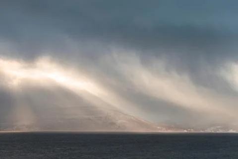 Dramatic sun rays break through storm clouds over a dark fjord in Northern .. Stock Photos