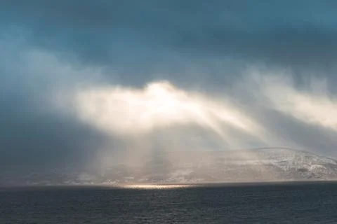 Dramatic sun rays break through storm clouds over a dark fjord in Northern .. Stock Photos
