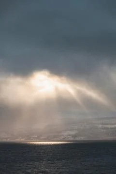 Dramatic sun rays break through storm clouds over a dark fjord in Northern .. Stockfoto's