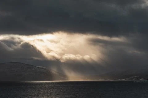 Dramatic sun rays break through storm clouds over a dark fjord in Northern .. Stock Photos