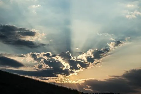 Dramatic sun rays pierce through clouds at sunset over a silhouetted landscap Stock Photos