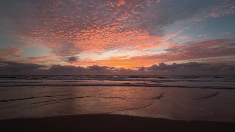 Dramatic sunrise over ocean waves with fiery clouds and wet sand reflections. 库存影片 333136928