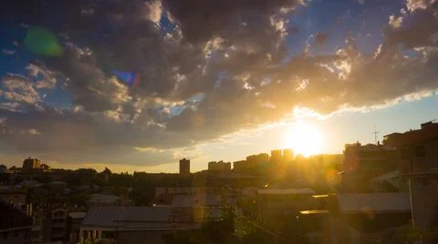 Dramatic sunset over cityscape with gray fluffy clouds and golden sun in blue Stock Photos