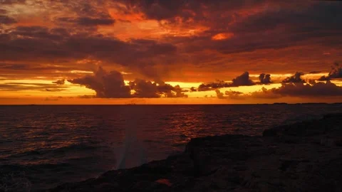 Dramatic Sunset Over Ocean with Waves Crashing Against Rocky Cliff and Fiery Sky Stock Footage 331850784