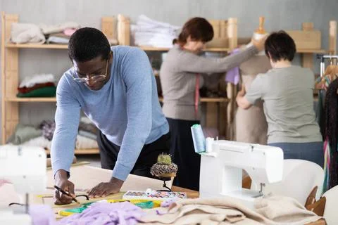 Dressmaker sketching sewing pattern while colleagues adjusting dress on Stock Photos