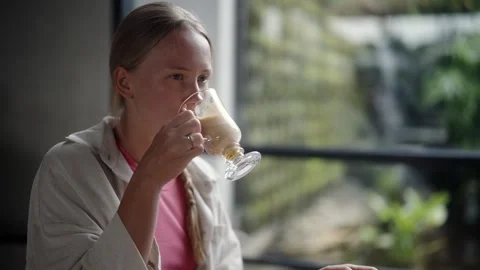 Drinking latte coffee in restaurant, sitting by the window. Woman in coffeeshop Stock Footage 296488113