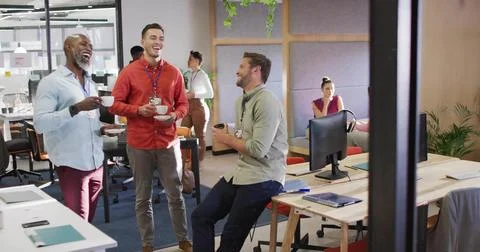 Drinking mugs of coffee, three men leaning around desk in modern office with  Stock Photos