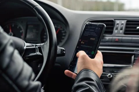 A driver in a black leather jacket is texting on a smartphone behind the whee Stock Photos