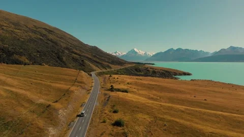 Drone aerial above van driving on road towards iconic mountain range & lake Stock Footage