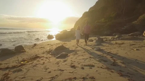 Drone aerial pull out of young lovers walking on beach with sunset Stock Footage 83929191