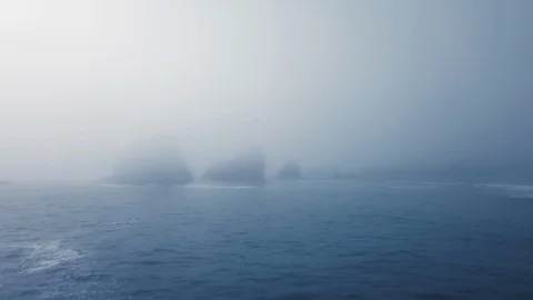 Drone camera approaches the rocks in dense fog protruding from the blue water Video stock 123392432