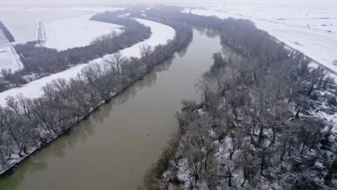 Drone capturing a river flanked by bare trees and snow-covered terrain in winter Stock Footage 263359073