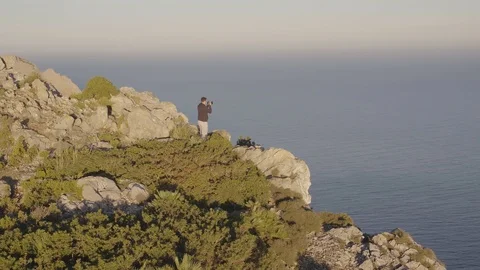 Drone circling guy with camera on cliff top going up Stock Footage 101427766
