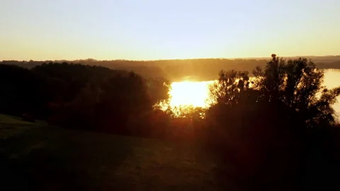 A drone flies from a field over trees towards the sun over a lake in which the Stock Footage 239101963