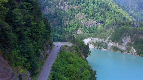 Drone flies forward above Enguri Reservoir road beside turquoise water and Vídeo Stock 343543051