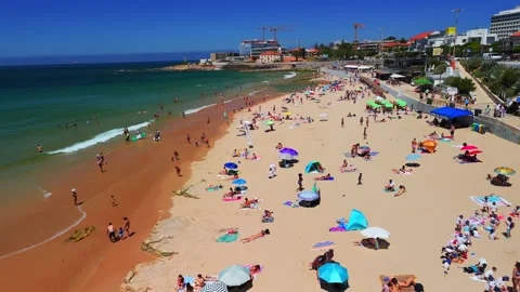 A drone flies over Carcavelos Beach with umbrellas, swimmers, and seafront road Видео 331066655