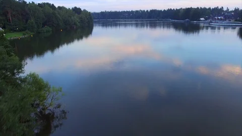The drone flies over the lake in evening among the trees, houses are on the sho Stock Footage 128698078