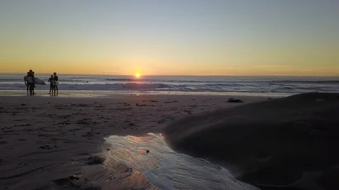 Drone flies over sand beach at sunset with surfers profiled in the distance 库存影片 100867275