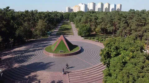 A drone flies over a square and follows cyclists passing by an green pyramid Stock Footage