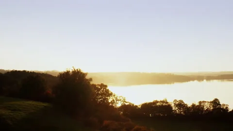 A drone flies towards the sun over the treetops to a lake. It's foggy during the 스톡 동영상 239100583