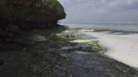 Drone flies under cliff overhanging onto sand and tide pools on a sunny day Stock Footage 344057119