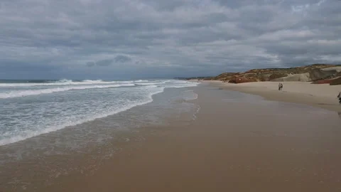 Drone flight over sandy beach with ocean splashing on overcast day in Nazare Stock Footage 157114219