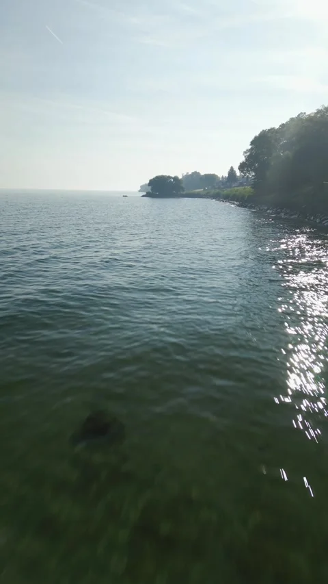 Drone flying fast over calm water toward shoreline with sparkling reflections Video stock 317279234
