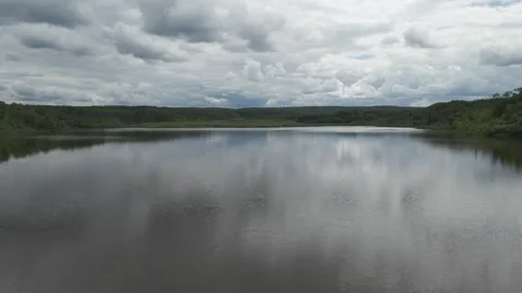 Drone flying low over a small lake surrounded by green trees under cloud filled Stock Footage 306622963