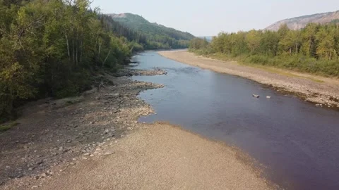 Drone flying over shallow river with rocky shoreline with large hills and trees Stock Footage 252779145