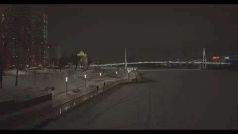 Drone flying over snowy river embankment with illuminated bridge at night Stock Footage 320304733