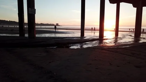 Drone glides 3 feet over beach with sunset seen through pier pillars 库存影片 85454009