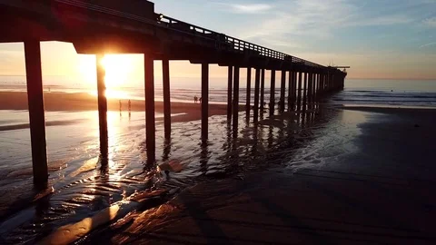 Drone glides beach to water edge with setting sun seen through pier pillars Stock Footage 85453489