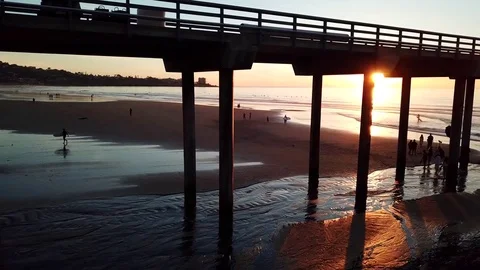 Drone glides beach to water edge with sunset seen through pier pillars Stock Footage 85453889