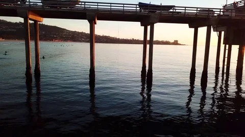 Drone glides fast 10 feet over water along pier with surfers catching waves 库存影片 85453583