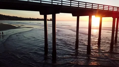 Drone glides water to beach with sunset seen through pier pillars 库存影片 85453978