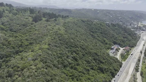 Drone lowers over forested hill next to coastal highway below paragliders Stock Footage 326769809