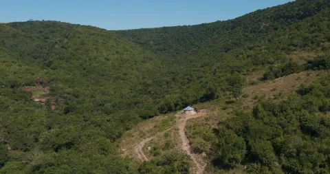 A drone moves over hills to a pathway leading to a rural hut in a clearing Stock Footage 242992778