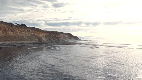 Drone pass along a rugged cliff shoreline with dark sand and rolling surf Stock Footage 330991709
