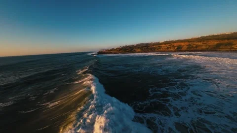 Drone shot follows waves breaking near rugged coastline at golden hour Stock Footage 339788988