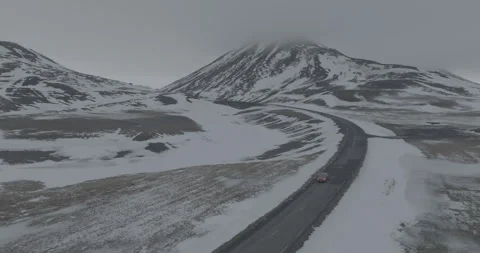 Drone Shot of Red Car Moving on Wet Road in Snowy Landscape of Iceland on Stock Footage 241081180
