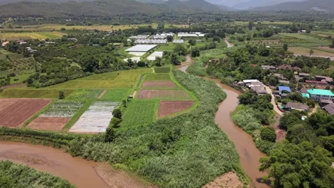 Drone slow tracking shot over muddy river bend, crop fields and greenhouses Stock Footage 335806130
