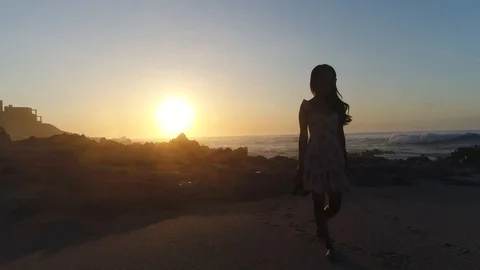 Drone tracking female model barefoot walking along beach at sunset backlit Stock Footage 104888145