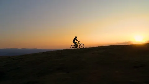 DRONE: Unrecognizable cyclist rides an electric bike uphill at golden sunset. Stock Footage