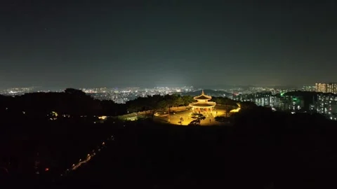Drone view of distant pavilion glowing above forest with city skyline at night Stock Footage 313641521