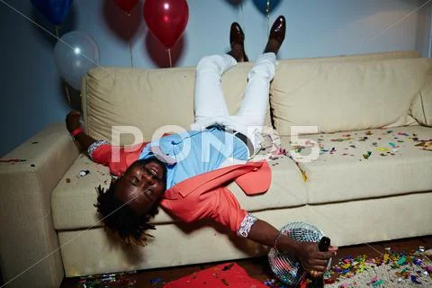 Drunk guy with bottle of champagne lying on sofa in messy room after party Stock Photos