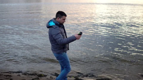 A drunk man in a jacket walks along the river bank and throws a can into the Stock Footage 142895142