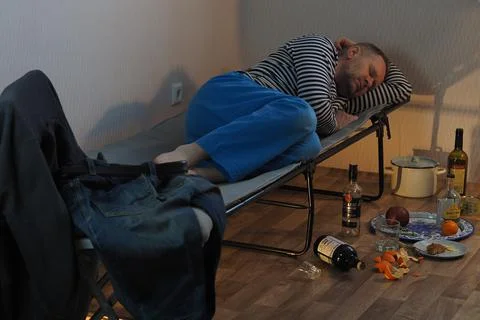 A drunk man lies on a cot and an empty bottle of alcohol is scattered nearby. Stock Photos