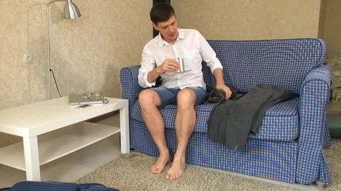 A drunk man, sitting on the couch, drinking alcohol, stroking a gray cat Stock Footage 86134675