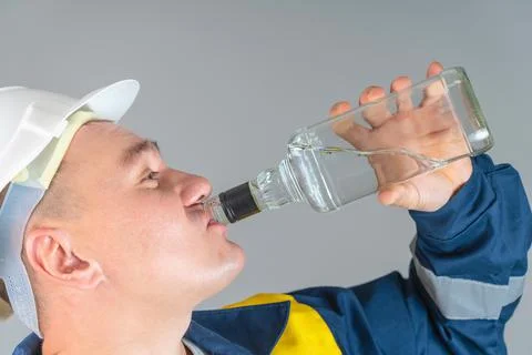 A drunk worker in a helmet greedily drinks strong alcohol from the throat of  Stock Photos